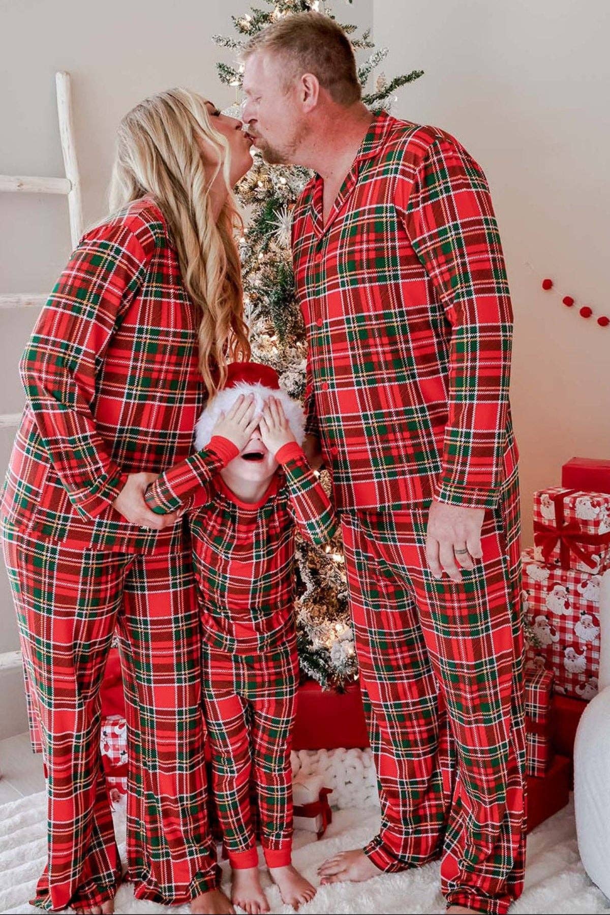 A family of four wearing matching red, green, and black plaid Christmas pajamas, standing together in front of a decorated Christmas tree and wrapped gifts.
