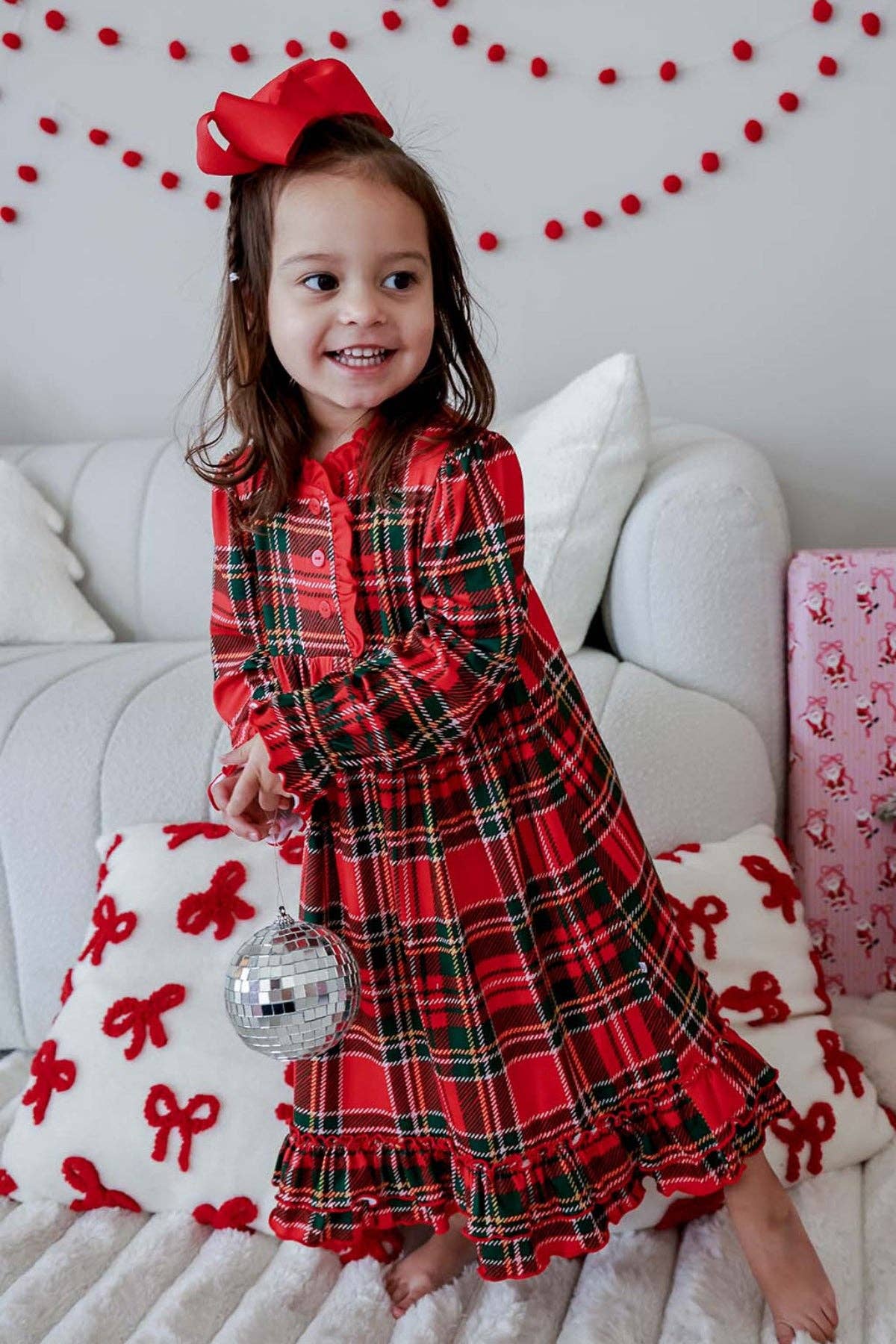 A young girl wearing a festive red and black plaid holiday dress with a ruffled hem, sitting on a white sofa decorated with red bows and a pink gift box. She is holding a silver disco ball accessory and has a red bow in her hair.