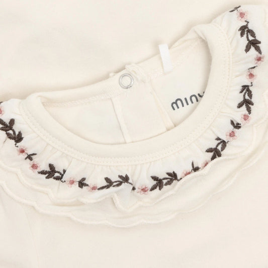 Close-up of a cream-colored baby top featuring a ruffled collar with dark brown leaf embroidery and small pink floral accents. The brand name 'minu' is partially visible on the inner neck tag.