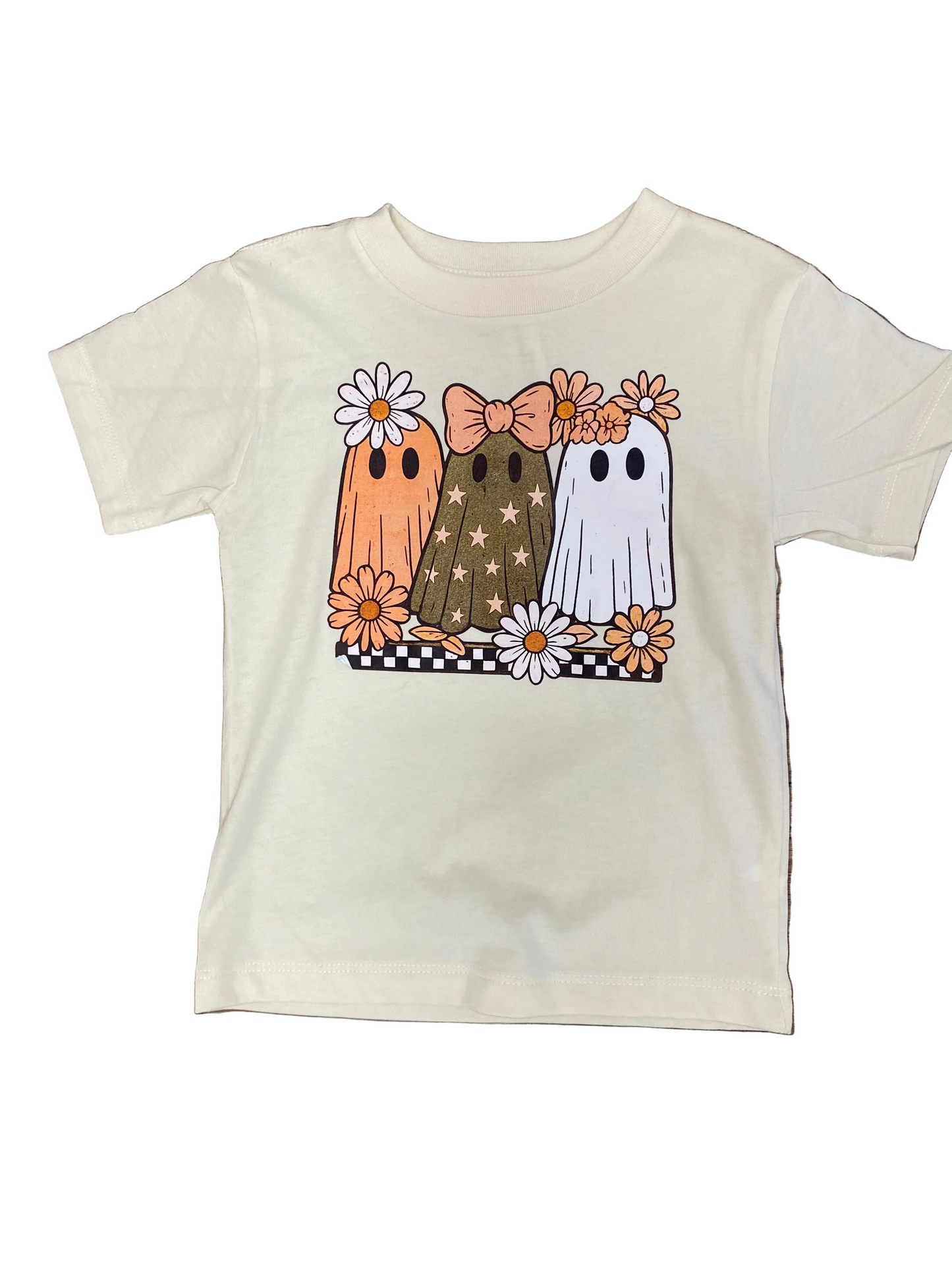 An off-white short-sleeve kids' t-shirt laid flat on a brown wood floor, featuring a graphic of three cute ghosts in orange, brown, and white with flowers.