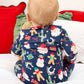 A baby seen from behind wearing a navy blue Christmas-themed footed sleeper with a print of Santa Claus, gingerbread men, snowmen, cacti, and gifts, sitting on a white bed with red and green pillows.