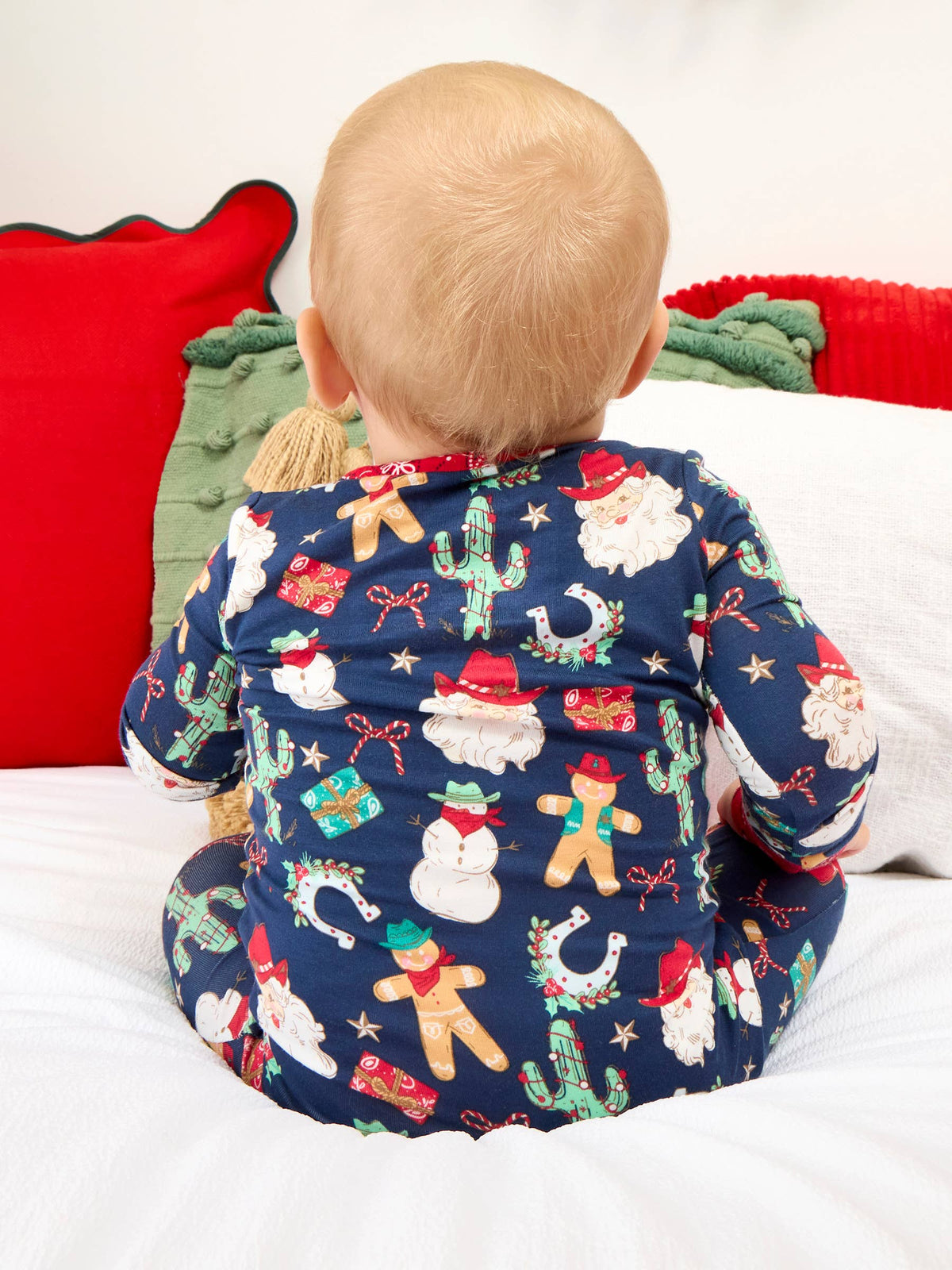A baby seen from behind wearing a navy blue Christmas-themed footed sleeper with a print of Santa Claus, gingerbread men, snowmen, cacti, and gifts, sitting on a white bed with red and green pillows.