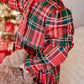 A child wearing a red and green plaid long-sleeve dress with ruffled trim, holding a brown teddy bear near a decorated Christmas tree.