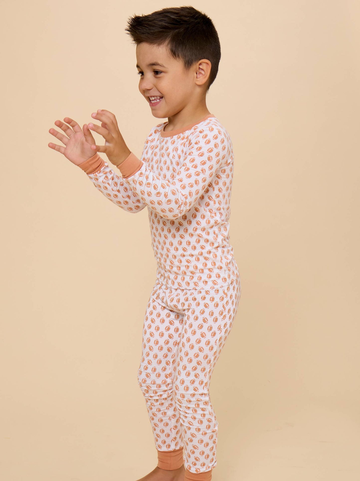 A young boy wearing a long-sleeve, one-piece pajama set with a white base and a repeating peach-orange heart pattern, standing against a solid beige studio background.