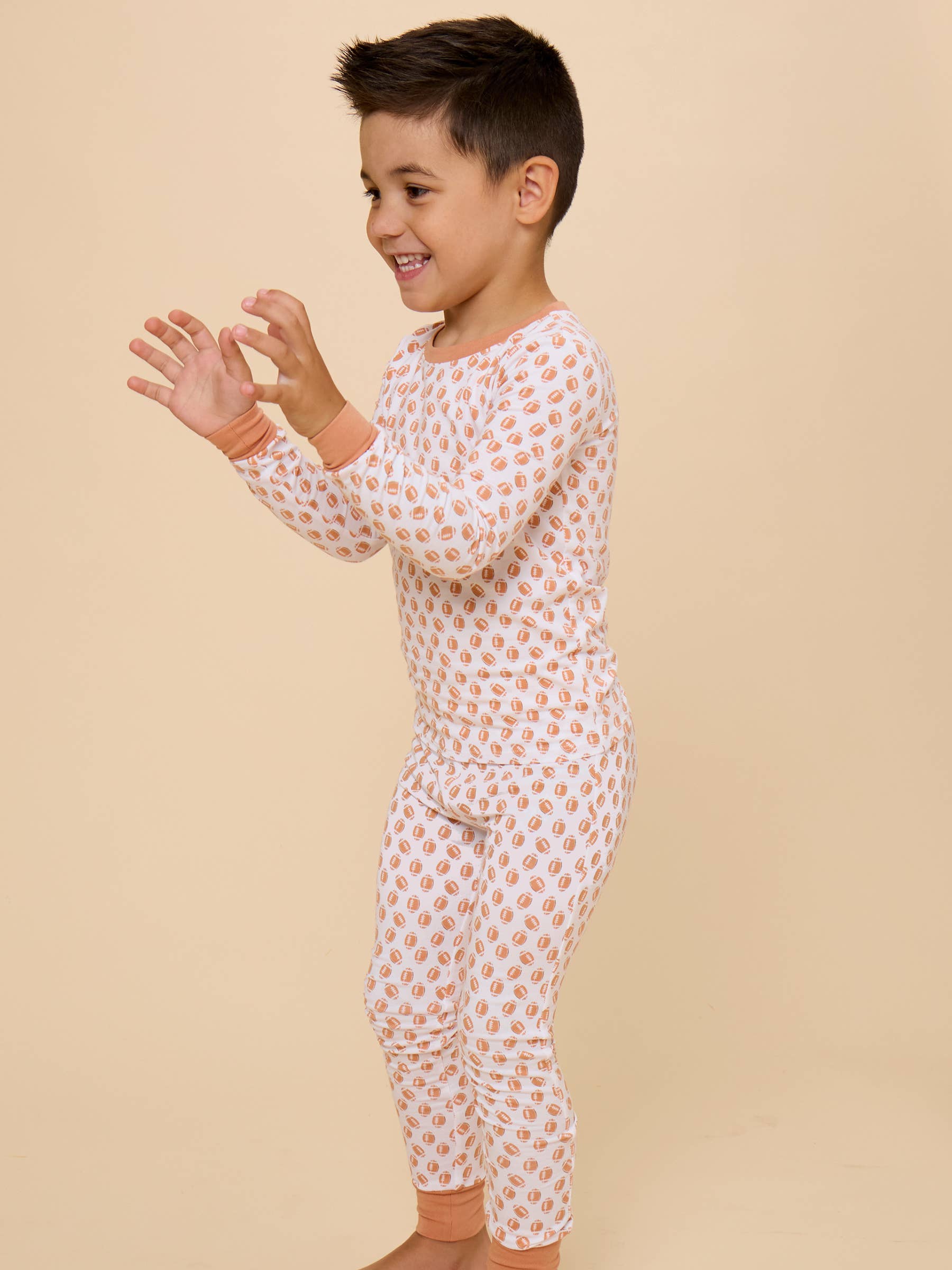 A young boy wearing a long-sleeve, one-piece pajama set with a white base and a repeating peach-orange heart pattern, standing against a solid beige studio background.