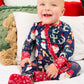 A smiling baby sitting on a white bed wearing a navy blue Christmas-themed footed sleeper with red trim and a Santa Claus and holiday print.