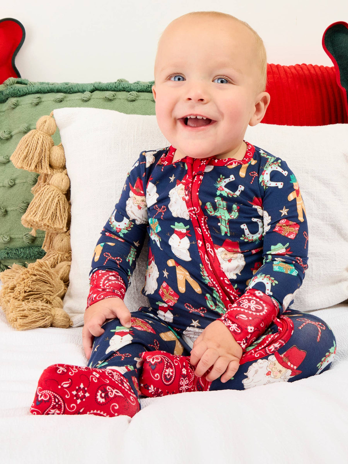 A smiling baby sitting on a white bed wearing a navy blue Christmas-themed footed sleeper with red trim and a Santa Claus and holiday print.