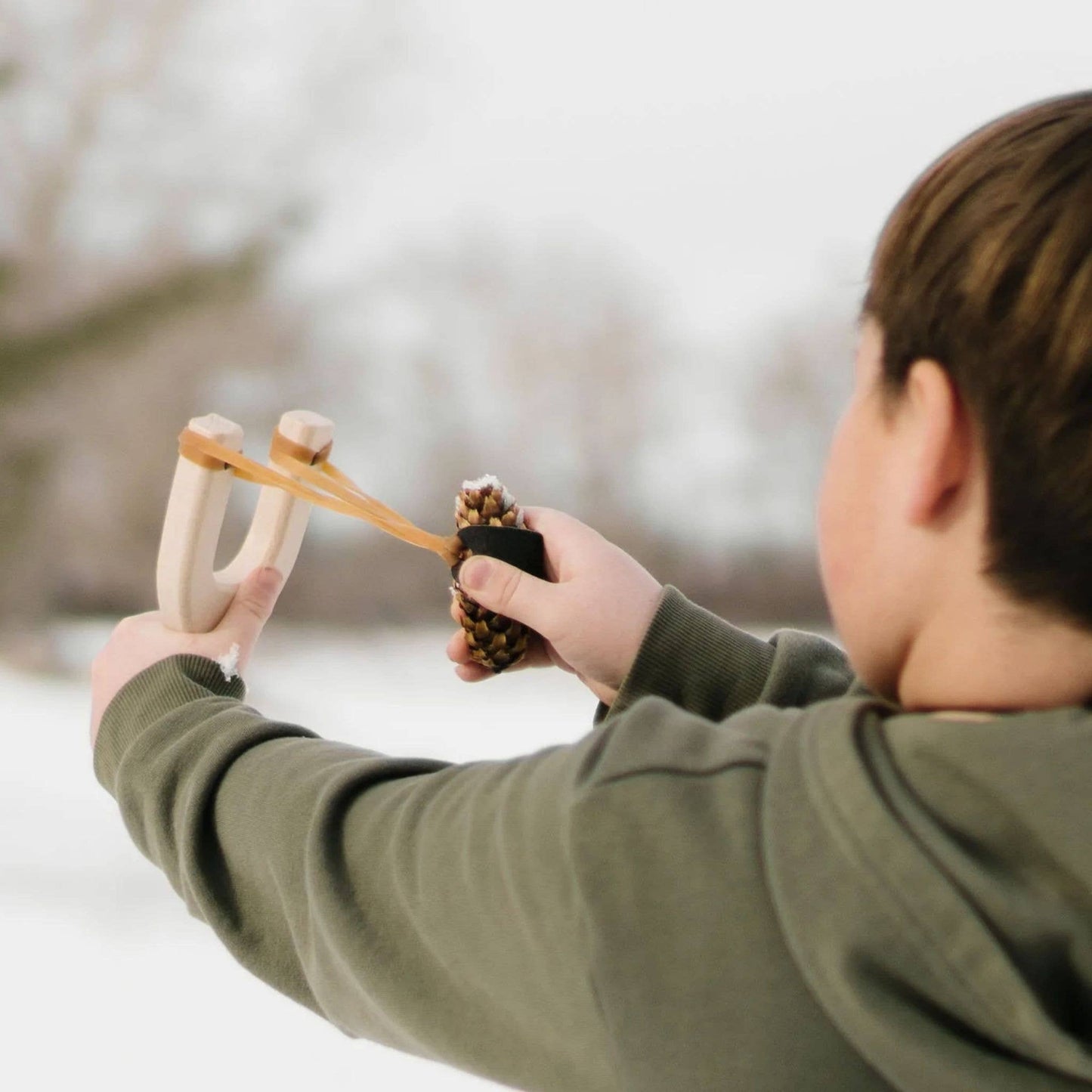 Child in a green jacket using a slingshot with a pinecone in a snowy landscape