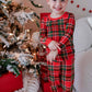 A young child wearing a red and green plaid long-sleeve pajama set with matching pants, standing barefoot in a cozy room next to a decorated Christmas tree and wrapped gifts.