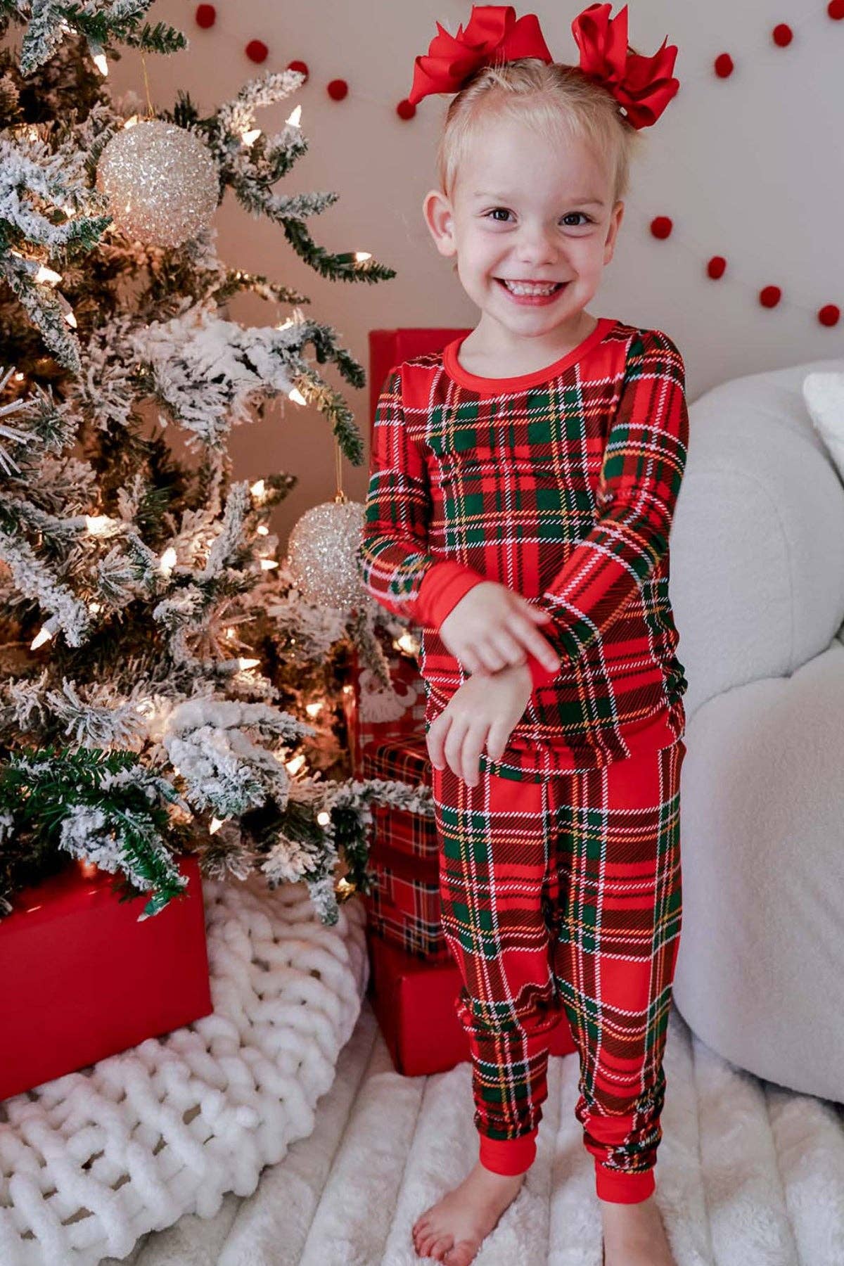 A young child wearing a red and green plaid long-sleeve pajama set with matching pants, standing barefoot in a cozy room next to a decorated Christmas tree and wrapped gifts.