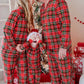 A family of four wearing matching red, green, and black plaid Christmas pajamas, standing together in front of a decorated Christmas tree and wrapped gifts.