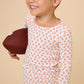 A smiling young boy wearing a white long-sleeve pajama top with a peach football print, holding a brown football against a solid beige background.
