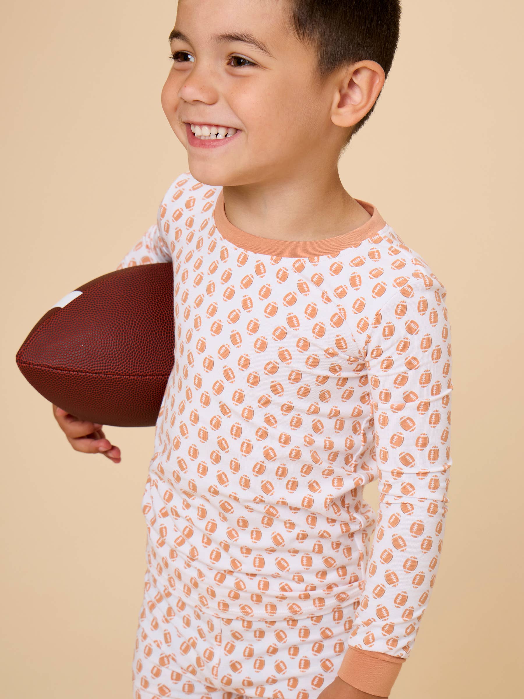 A smiling young boy wearing a white long-sleeve pajama top with a peach football print, holding a brown football against a solid beige background.