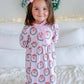 A young child wearing a pink holiday-themed long-sleeve pajama romper with a snowman and wreath pattern, standing on a white quilted bed with pillows and a festive wreath in the background.