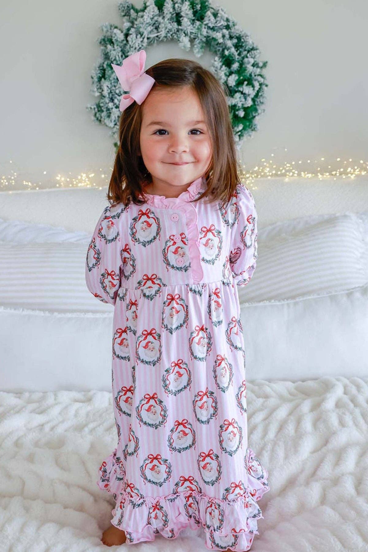 A young child wearing a pink holiday-themed long-sleeve pajama romper with a snowman and wreath pattern, standing on a white quilted bed with pillows and a festive wreath in the background.