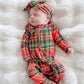A sleeping newborn baby wearing a red and green tartan plaid footed one-piece jumpsuit with a matching plaid bow headband, lying on a white faux fur blanket.