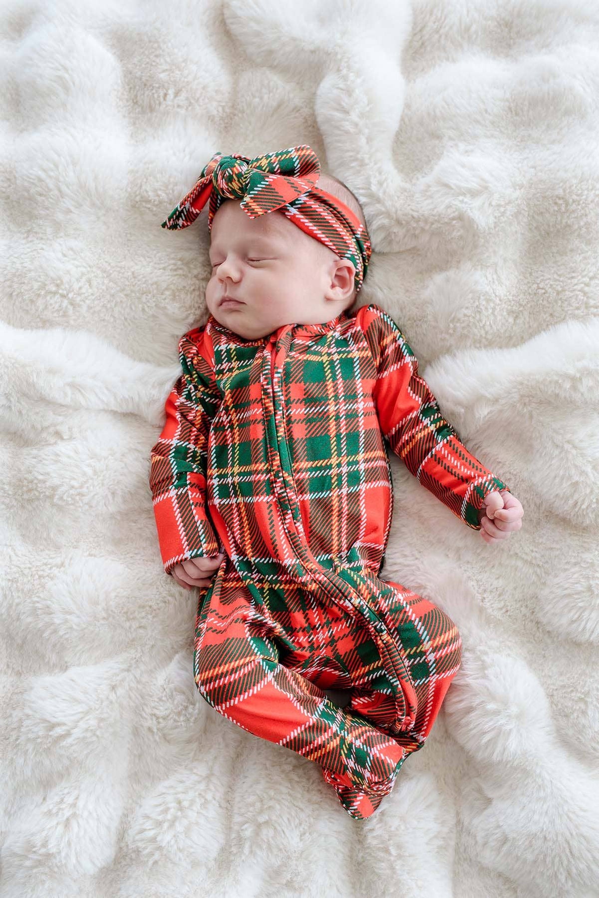 A sleeping newborn baby wearing a red and green tartan plaid footed one-piece jumpsuit with a matching plaid bow headband, lying on a white faux fur blanket.