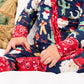 A close-up of a child wearing a navy blue and red Christmas-themed long-sleeve pajama romper with festive prints, sitting on a white cushion.