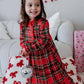 A young girl wearing a festive red and black plaid holiday dress with a ruffled hem, sitting on a white sofa decorated with red bows and a pink gift box. She is holding a silver disco ball accessory and has a red bow in her hair.