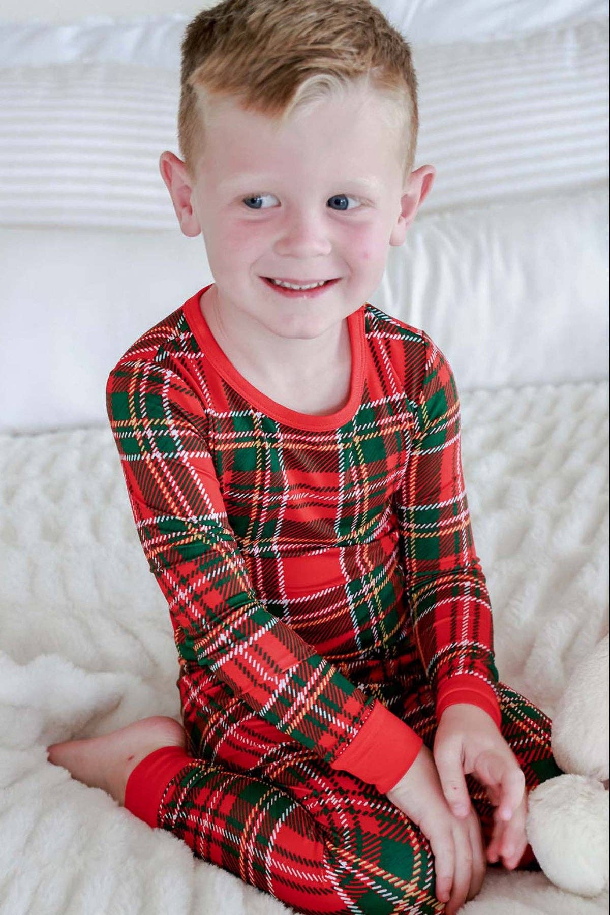 A young boy smiling while wearing a red and green plaid long-sleeve pajama set, sitting on a white textured blanket.