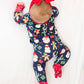 A baby lying on a white fabric surface wearing a navy blue Christmas-themed footed onesie with a red bow headband and red mittens.