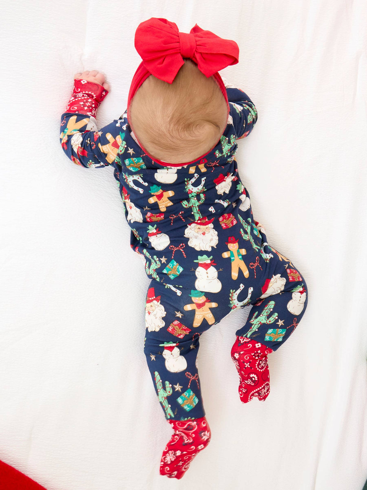 A baby lying on a white fabric surface wearing a navy blue Christmas-themed footed onesie with a red bow headband and red mittens.