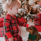 A toddler wearing a red, green, and black plaid Christmas pajama set kneels on a white blanket next to a brown dachshund dog with a green bow tie, in front of a decorated Christmas tree and wrapped gifts.