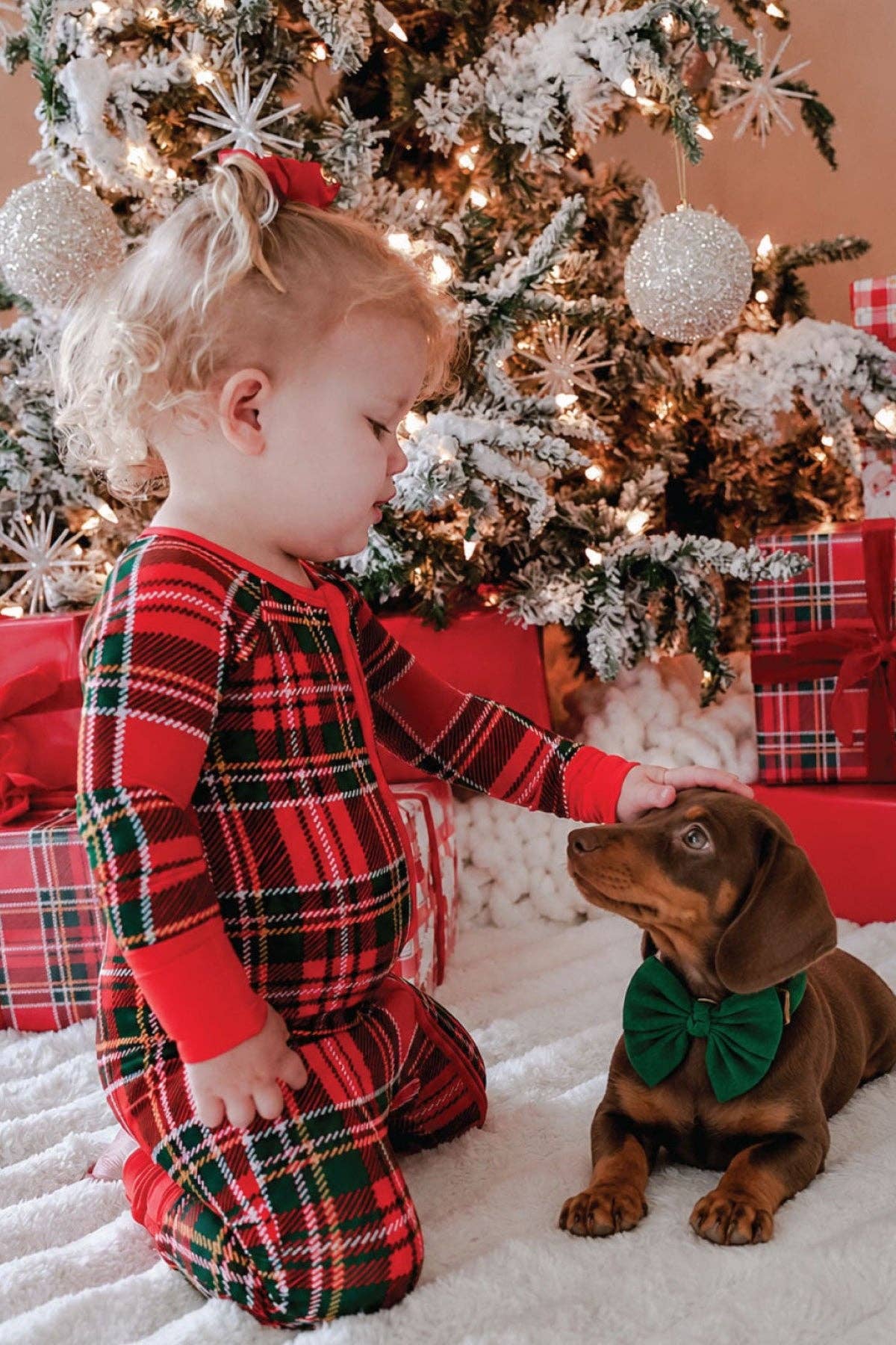 A toddler wearing a red, green, and black plaid Christmas pajama set kneels on a white blanket next to a brown dachshund dog with a green bow tie, in front of a decorated Christmas tree and wrapped gifts.