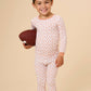 A young boy smiling and holding a brown football, wearing a long-sleeve pajama set with a white base and a repeating orange-brown pattern, set against a solid beige studio background.