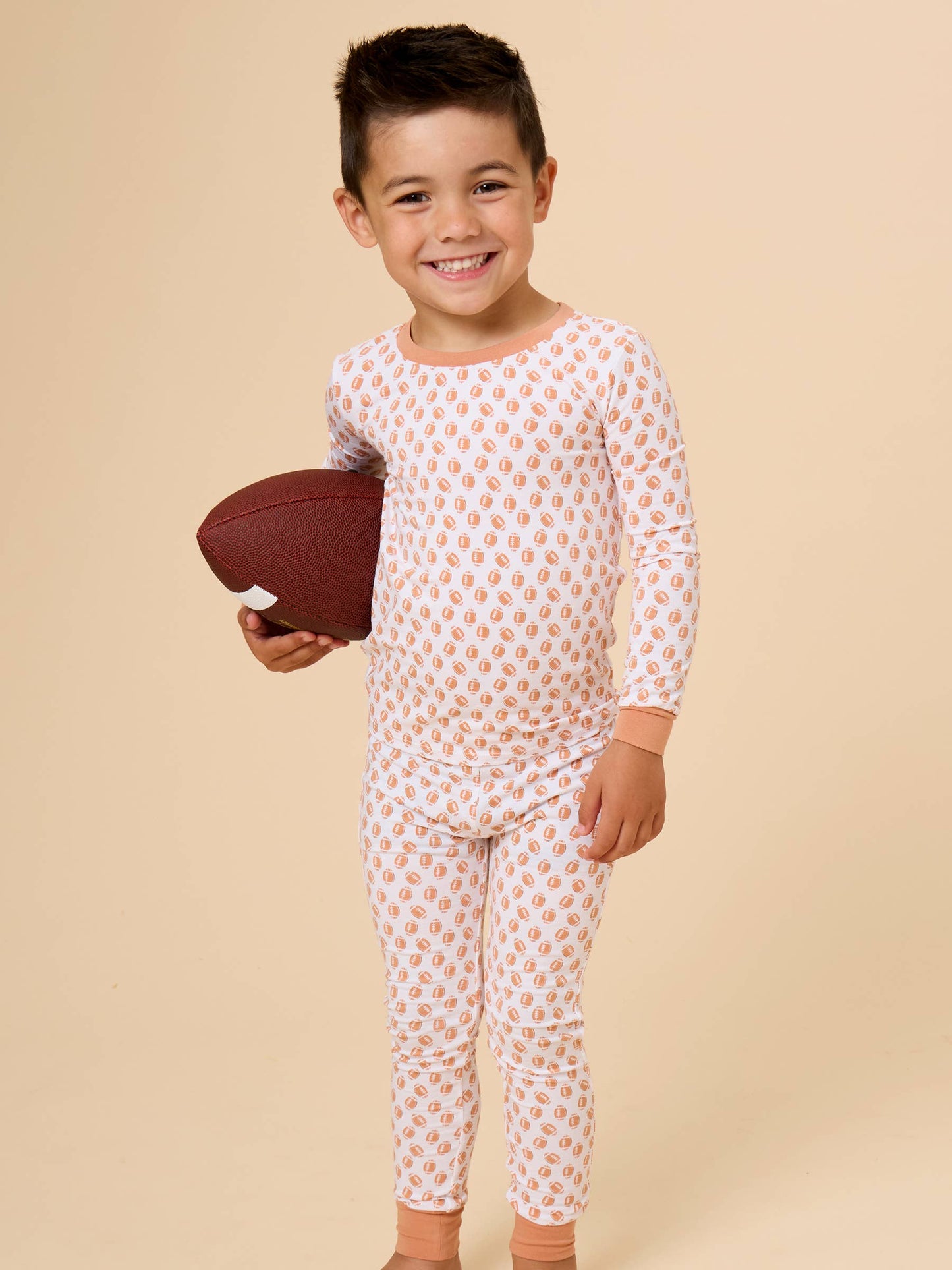 A young boy smiling and holding a brown football, wearing a long-sleeve pajama set with a white base and a repeating orange-brown pattern, set against a solid beige studio background.