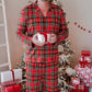 A man wearing a red and green plaid pajama set stands barefoot in front of a decorated Christmas tree with wrapped gifts, holding a small white gift box.
