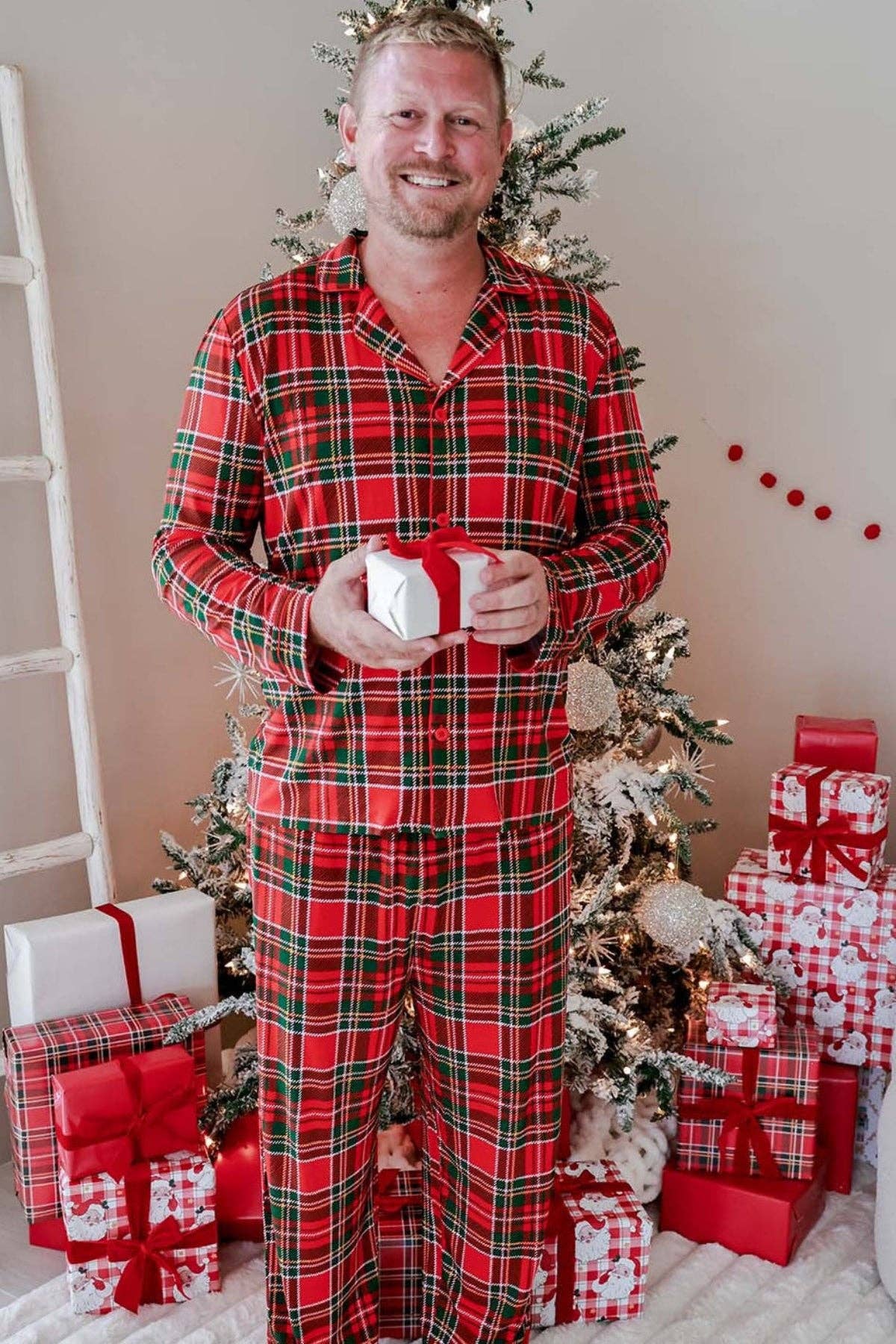 A man wearing a red and green plaid pajama set stands barefoot in front of a decorated Christmas tree with wrapped gifts, holding a small white gift box.