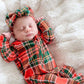 A sleeping newborn baby wearing a red, green, and black plaid footed onesie with a matching plaid bow headband, lying on a soft white faux fur blanket.