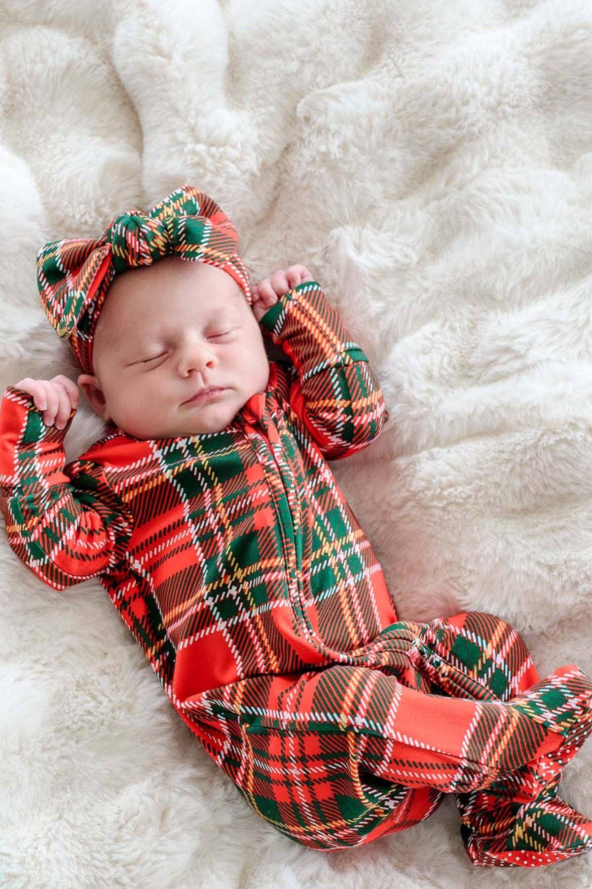 A sleeping newborn baby wearing a red, green, and black plaid footed onesie with a matching plaid bow headband, lying on a soft white faux fur blanket.