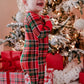 A toddler girl wearing a red, green, and black plaid Christmas pajama romper stands smiling in front of a decorated Christmas tree with wrapped gifts and white fluffy decorations.