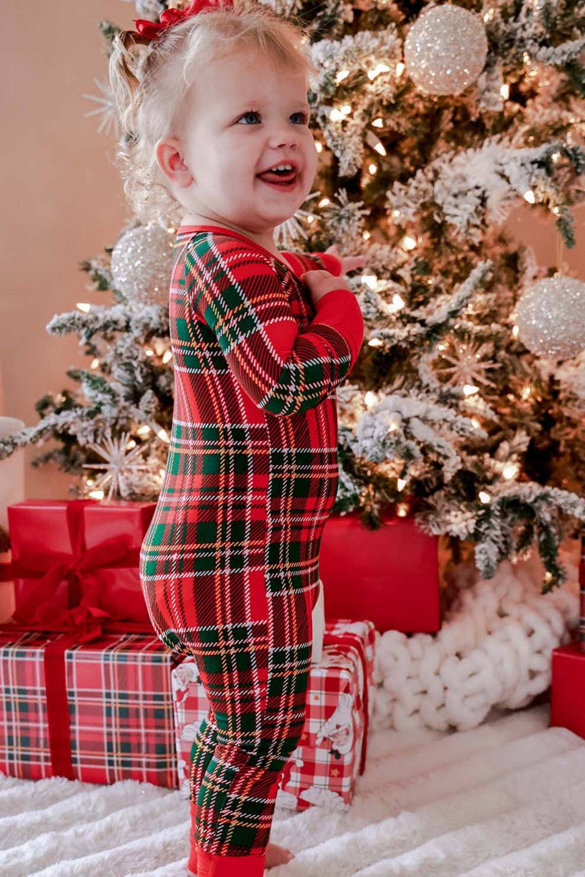 A toddler girl wearing a red, green, and black plaid Christmas pajama romper stands smiling in front of a decorated Christmas tree with wrapped gifts and white fluffy decorations.