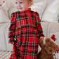 A toddler girl wearing a red and green plaid long-sleeve dress with a matching red bow in her hair, standing next to a brown teddy bear in front of a white couch.