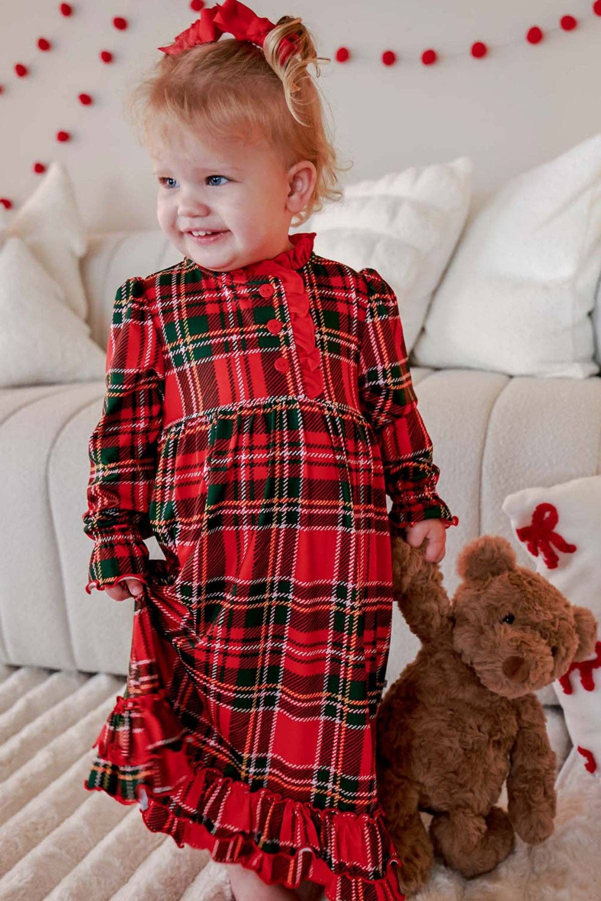 A toddler girl wearing a red and green plaid long-sleeve dress with a matching red bow in her hair, standing next to a brown teddy bear in front of a white couch.