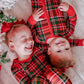 Two young children lying on a white faux-fur surface, wearing matching red holiday plaid zip-up pajamas with green and white tartan patterns. A decorated Christmas tree branch and red bow are visible at the left edge.