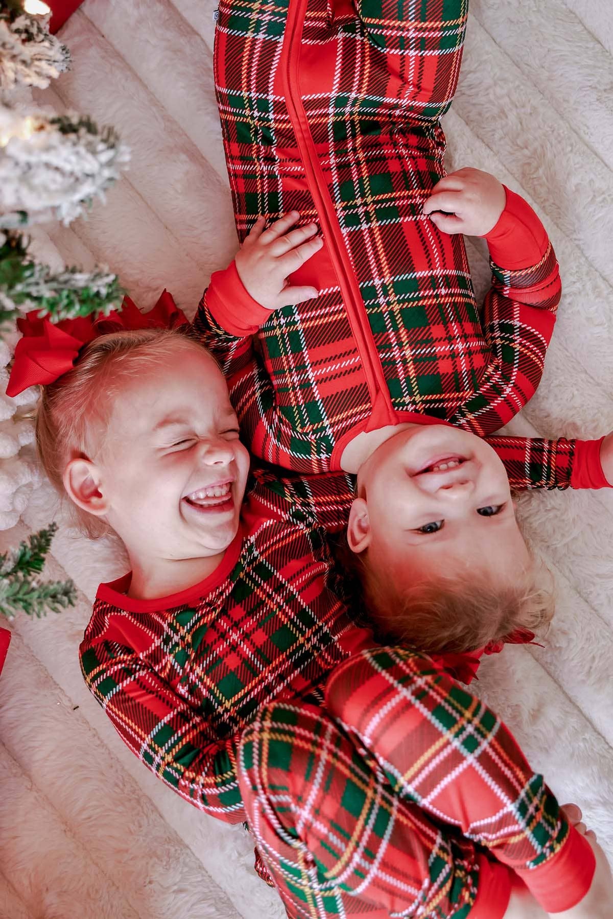 Two young children lying on a white faux-fur surface, wearing matching red holiday plaid zip-up pajamas with green and white tartan patterns. A decorated Christmas tree branch and red bow are visible at the left edge.