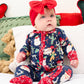 A baby wearing a festive navy blue Christmas-themed footed sleeper with red trim and a matching red bow headband, sitting on a white cushion with holiday pillows.