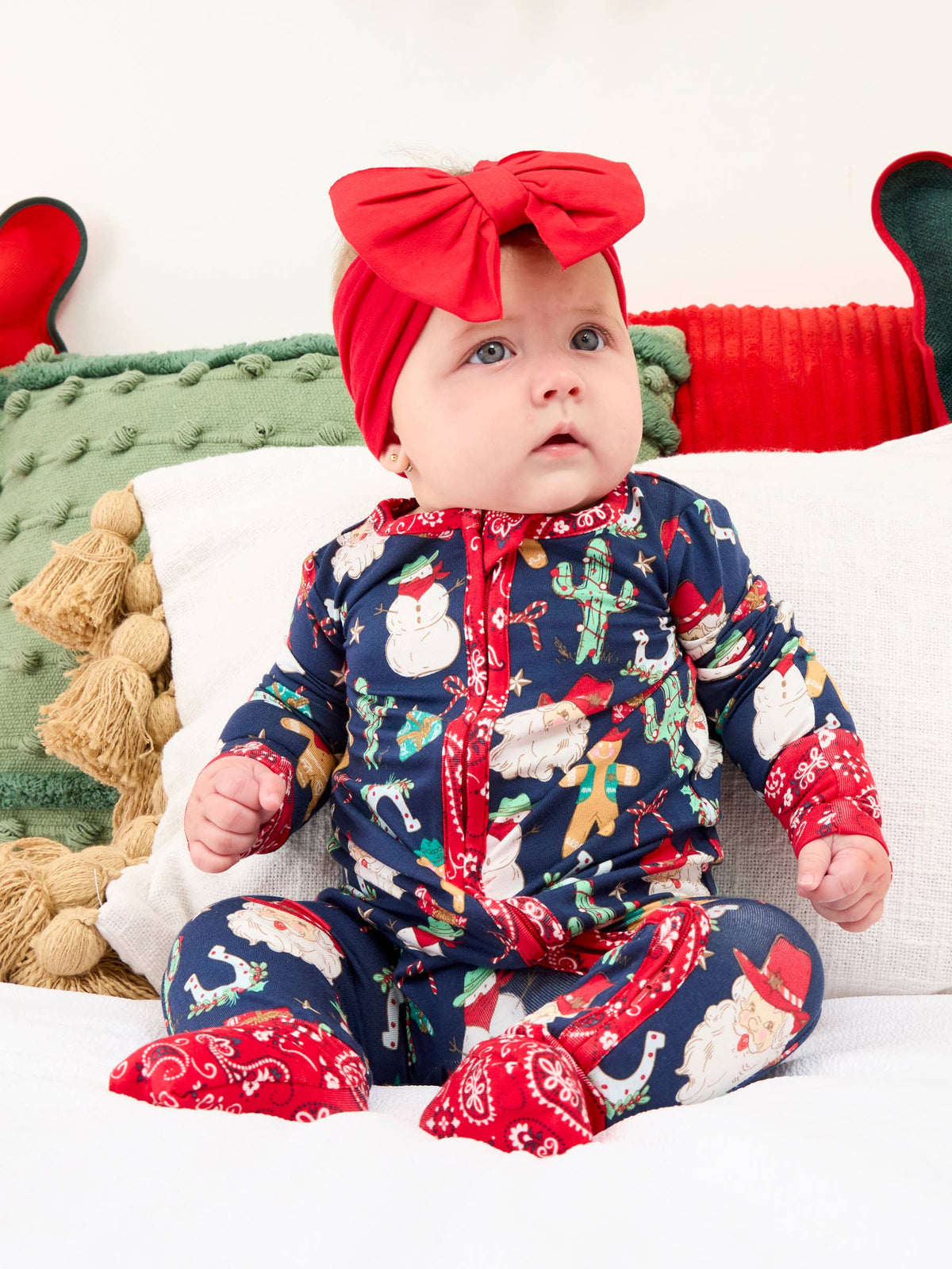 A baby wearing a festive navy blue Christmas-themed footed sleeper with red trim and a matching red bow headband, sitting on a white cushion with holiday pillows.
