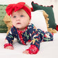 A baby lying on a white bed wearing a navy blue Christmas-themed long-sleeve footed sleeper with a red bow headband.
