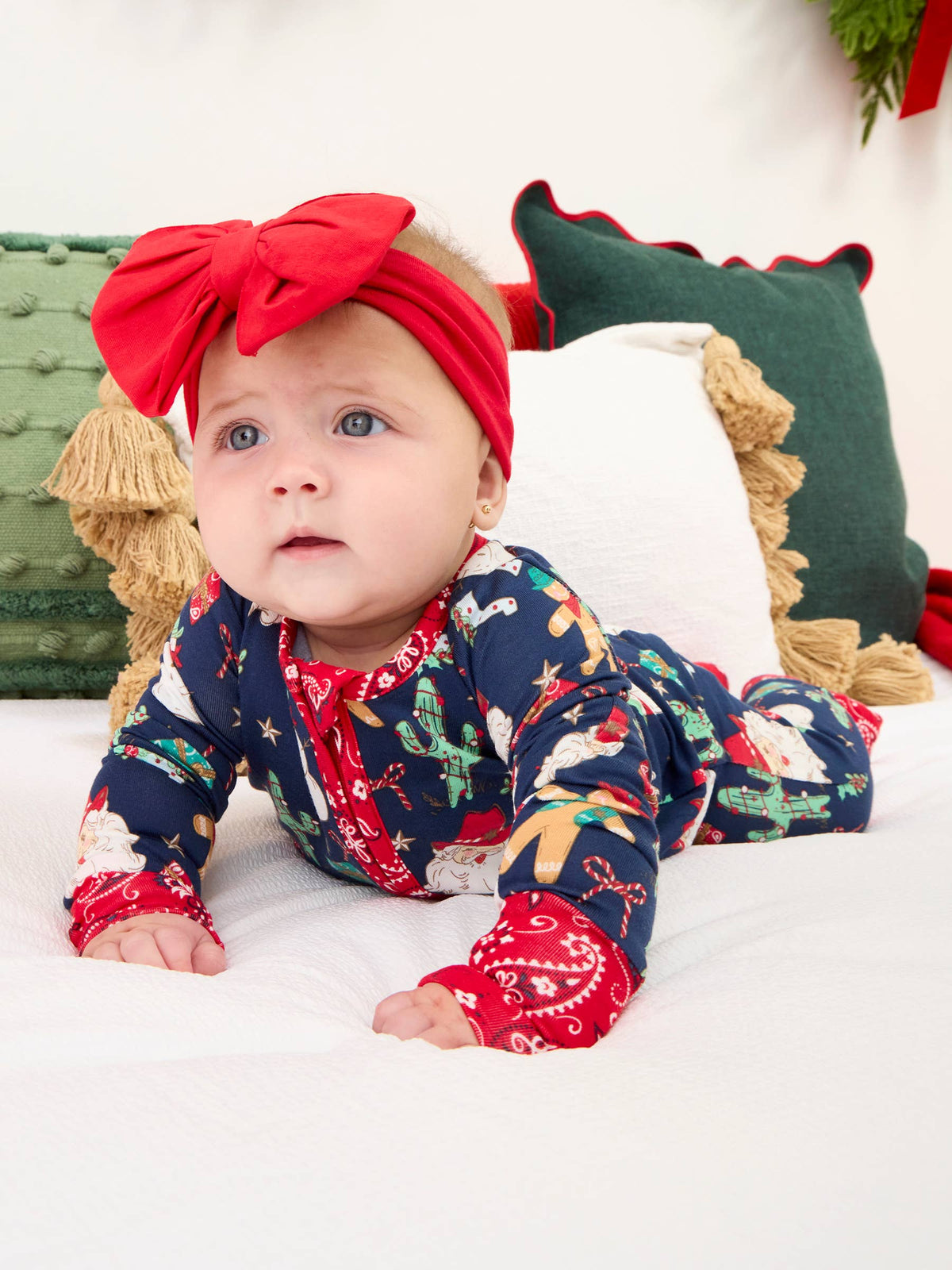 A baby lying on a white bed wearing a navy blue Christmas-themed long-sleeve footed sleeper with a red bow headband.