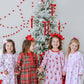 Four young girls wearing festive Christmas nightgowns stand barefoot in front of a decorated flocked Christmas tree. The girls are wearing matching holiday sleepwear in pink, red, and green patterns, including plaid and reindeer prints, with red bows in their hair.