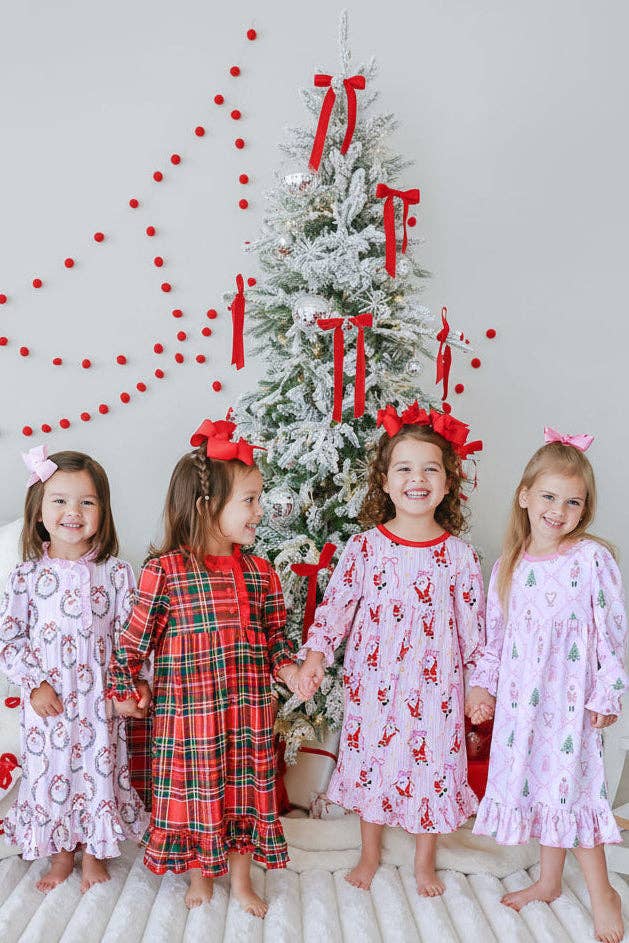 Four young girls wearing festive Christmas nightgowns stand barefoot in front of a decorated flocked Christmas tree. The girls are wearing matching holiday sleepwear in pink, red, and green patterns, including plaid and reindeer prints, with red bows in their hair.