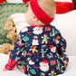 A baby wearing a navy blue hooded one-piece pajama with a festive Christmas print, featuring a red headband with a bow, sitting on a white bed.