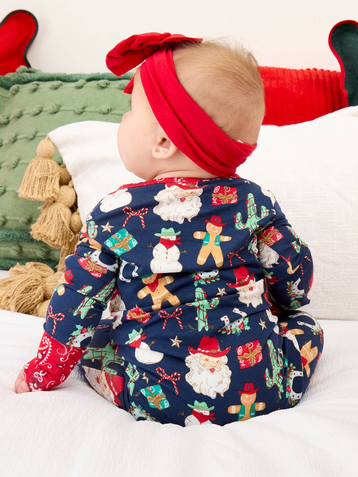 A baby wearing a navy blue hooded one-piece pajama with a festive Christmas print, featuring a red headband with a bow, sitting on a white bed.