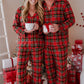 A smiling couple wearing matching red and green plaid pajamas stands barefoot in front of a decorated Christmas tree, each holding a white mug. Gift boxes are visible around them in a cozy indoor holiday setting.