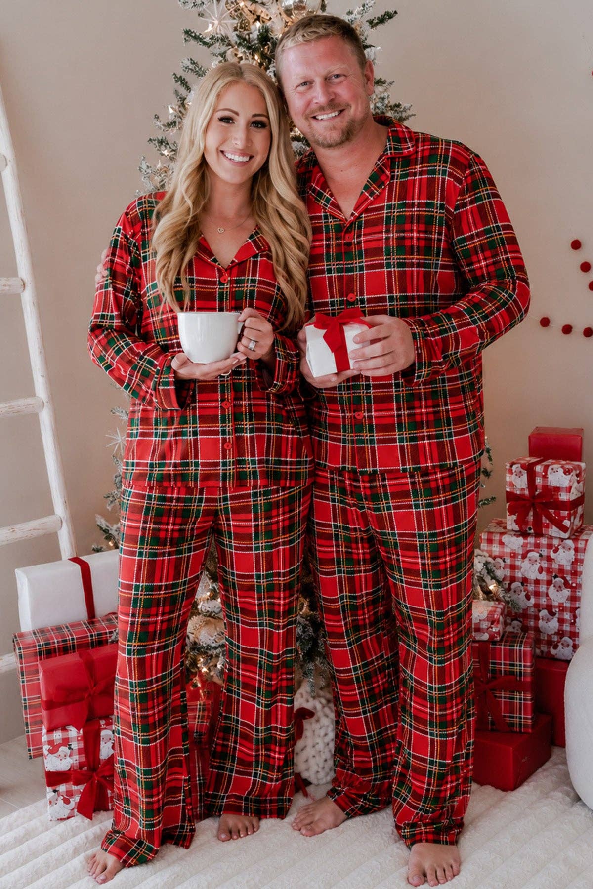 A smiling couple wearing matching red and green plaid pajamas stands barefoot in front of a decorated Christmas tree, each holding a white mug. Gift boxes are visible around them in a cozy indoor holiday setting.